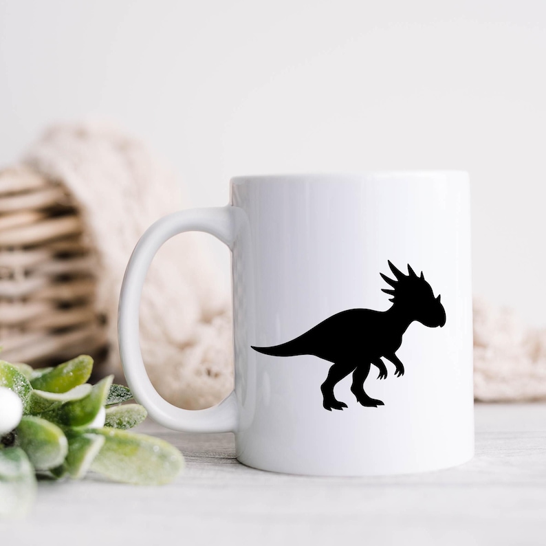 May include: White ceramic mug with a black silhouette of a Stygimoloch dinosaur. The dinosaur is shown in profile, with a spiky head and a long tail. The mug has a curved handle. The background is blurred.