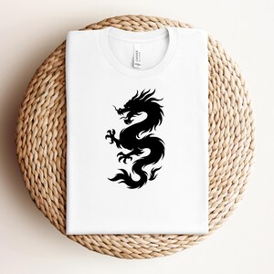 May include: A white t-shirt featuring a black dragon design. The dragon is a stylized silhouette, with intricate details. The shirt is displayed on a round, woven surface.