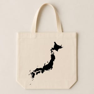 May include: A cream-colored tote bag featuring a black silhouette of the Japanese islands. The bag has two matching handles and is made of a sturdy fabric. The design is simple and graphic, perfect for carrying books, groceries, or personal items.