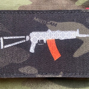 May include: A black patch with a white silhouette of an assault rifle with an orange magazine. The rifle is made of reflective material.