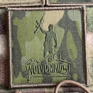 May include: A green, brown, and tan camouflage fabric patch with a dark green embroidered silhouette of a soldier holding a rifle. The text "WOLVERINES" is embroidered below the soldier in dark green.