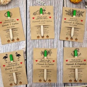 May include: A collection of kraft paper seed packets with green-topped wooden plant markers. Each packet features a unique graphic, the names "Hannah & Copelan", and the date "10.21.2026". The packets are arranged on a wooden surface.