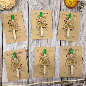 May include: Six pens with green tops and natural wood barrels are displayed on kraft paper cards. The cards feature floral illustrations and the text "LET LOG ROW" and "LIAM & OLIVIA" with a date. The pens are arranged on a wooden surface.