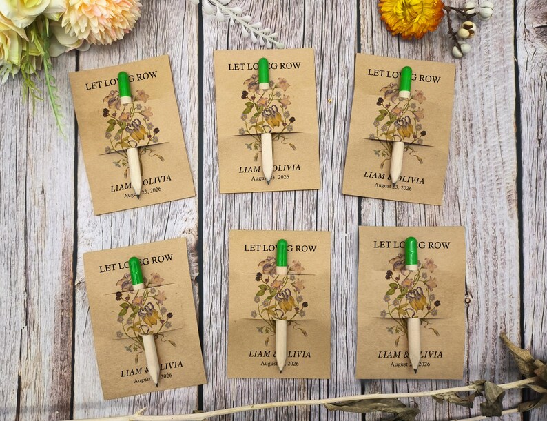 May include: Six pens with green tops and natural wood barrels are displayed on kraft paper cards. The cards feature floral illustrations and the text "LET LOG ROW" and "LIAM & OLIVIA" with a date. The pens are arranged on a wooden surface.