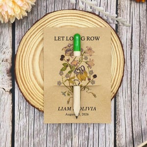 May include: A seed packet with the text "LET LOVE GROW" and the names "LIAM OLIVIA" and the date "August 3, 2026". The packet features a floral design and a green and white pencil. The packet is on a wooden slice.
