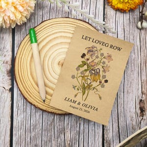 May include: A seed packet with the text "LET LOVEG ROW" and the names "LIAM & OLIVIA" with the date "August 23, 2026" printed on it. The packet features a floral design. A wooden pencil with a green tip rests on a wooden slice.