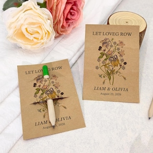 May include: Two kraft paper seed packets with floral illustrations and the text "LET LOVE GROW" and "LIAM & OLIVIA August 23, 2026". A pen with a green tip is inserted into one packet. Roses and a wood slice are in the background.