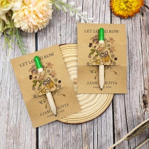 May include: Two seed packet favours with a pen. The kraft paper packets have the text "LET LOG ROW" and "LIAM OLIVIA August 3, 2026" printed on them. The packets feature a floral design and a pen with a green top.