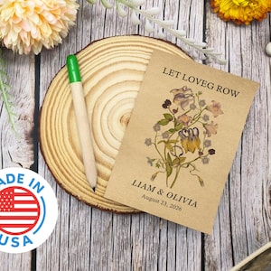 May include: A seed packet with the text "LET LOVE GROW" and the names "Liam & Olivia" with a date. The packet features a floral illustration and rests on a wooden slice with a green-tipped pencil. A "MADE IN USA" sticker is also visible.