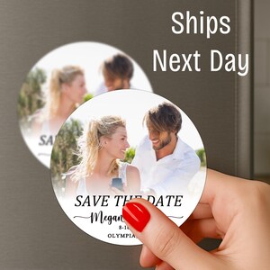 May include: Round magnet with a photo of a smiling couple, with the text "SAVE THE DATE" and a name. The magnet is held by a hand with red nail polish. The text "Ships Next Day" is also visible.