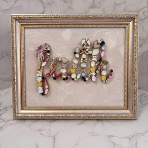 May include: A framed wall art piece featuring the word Faith spelled out in a collage of assorted vintage jewelry, including colorful rhinestones, pearls, and gold-tone metal accents, set against a textured cream background in a gold ornate frame.
