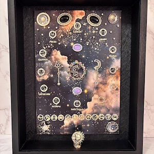 May include: A black shadow box frame containing a celestial-themed display with a dark nebula background. It features various gold-toned zodiac symbols, star-shaped pins, and decorative cabochons arranged around a central moon and sun motif.