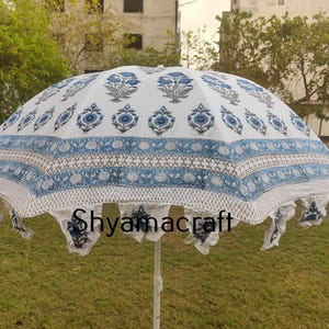 Puede incluir: Una sombrilla blanca y azul con estampado y borde de flecos. La sombrilla tiene un dise&ntilde;o floral con flores y hojas azules. El texto "Shyamacraft" est&aacute; impreso en la sombrilla.