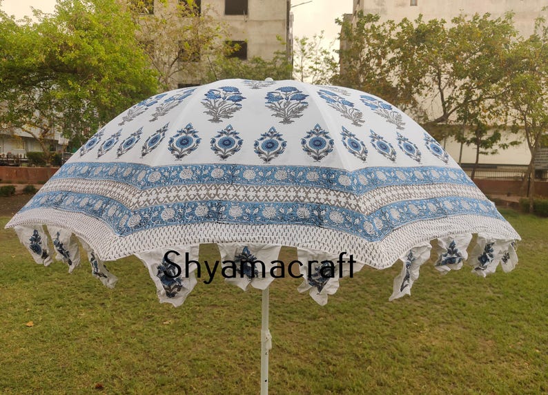 Puede incluir: Una sombrilla blanca y azul con estampado y borde de flecos. La sombrilla tiene un dise&ntilde;o floral con flores y hojas azules. El texto "Shyamacraft" est&aacute; impreso en la sombrilla.