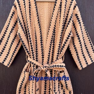 May include: A peach-coloured robe featuring a repeating black triangular pattern, long sleeves, a tie-waist belt, and two front pockets. The fabric has visible quilted stitching throughout, and the text Shyamacrafts is printed in blue.