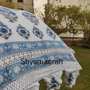 Puede incluir: Tela de algod&oacute;n blanca con estampado floral azul. La tela tiene un borde con un patr&oacute;n geom&eacute;trico azul y gris. La tela est&aacute; drapeada sobre una zona verde cubierta de hierba. El texto "Shyamacraft" es visible en la tela.