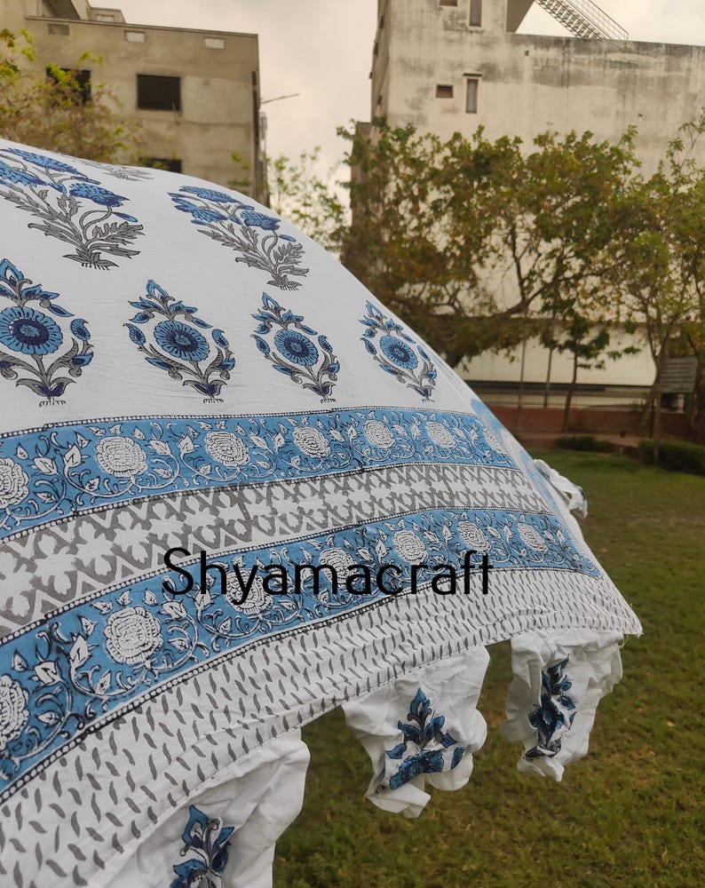 Puede incluir: Tela de algod&oacute;n blanca con estampado floral azul. La tela tiene un borde con un patr&oacute;n geom&eacute;trico azul y gris. La tela est&aacute; drapeada sobre una zona verde cubierta de hierba. El texto "Shyamacraft" es visible en la tela.
