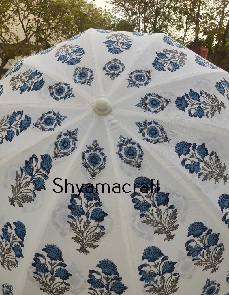 Puede incluir: Paraguas de algod&oacute;n blanco con estampado floral azul. El paraguas tiene un mango blanco y est&aacute; decorado con un patr&oacute;n repetitivo de flores y hojas azules. El texto "Shyamacraft" est&aacute; impreso en el centro del paraguas.