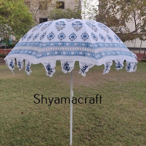 Puede incluir: Una sombrilla blanca y azul con estampado y un borde de flecos blancos. La sombrilla est&aacute; abierta y de pie sobre un c&eacute;sped. El texto "Shyamacraft" es visible en el poste.