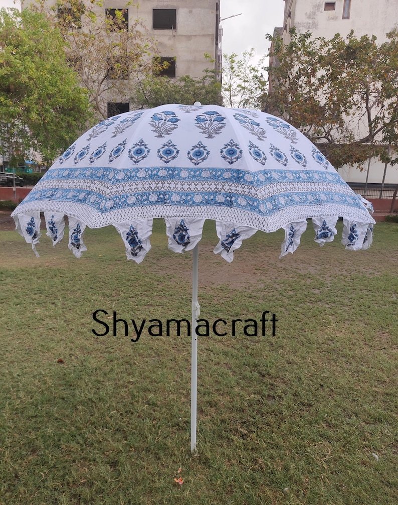Puede incluir: Una sombrilla blanca y azul con estampado y un borde de flecos blancos. La sombrilla est&aacute; abierta y de pie sobre un c&eacute;sped. El texto "Shyamacraft" es visible en el poste.