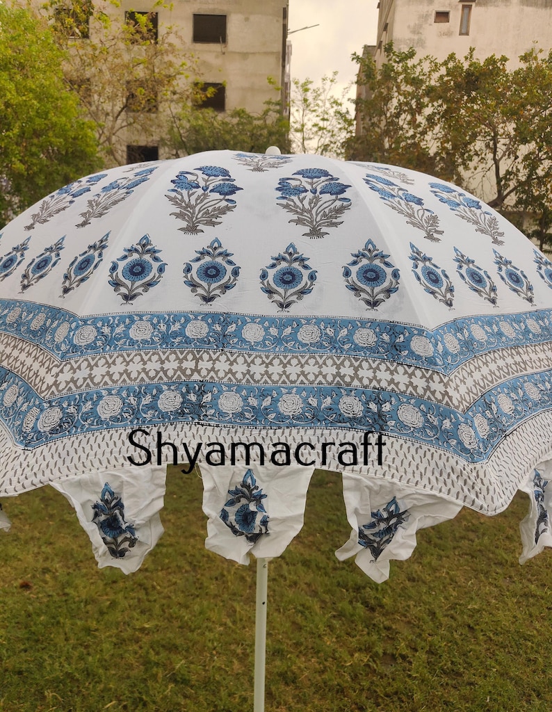 Puede incluir: Paraguas de algod&oacute;n blanco con estampado floral azul y gris. El paraguas tiene un fleco blanco alrededor del borde y las palabras "Shyamacraft" impresas en la tela.