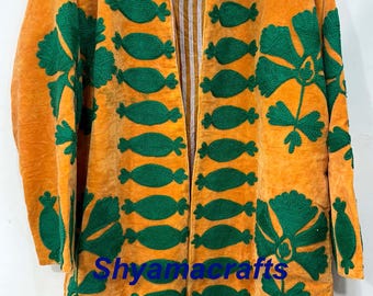 May include: An open-front, long-sleeved coat in a vibrant orange hue, adorned with intricate green floral embroidery. The coat features a striped interior lining and the text "Shyamacrafts" at the bottom. The design is a blend of color and texture.