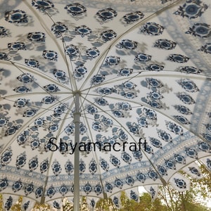Puede incluir: Una sombrilla blanca y azul con un poste plateado. La sombrilla tiene un dise&ntilde;o floral con flores y hojas azules sobre un fondo blanco. La sombrilla est&aacute; abierta y la tela est&aacute; estirada.