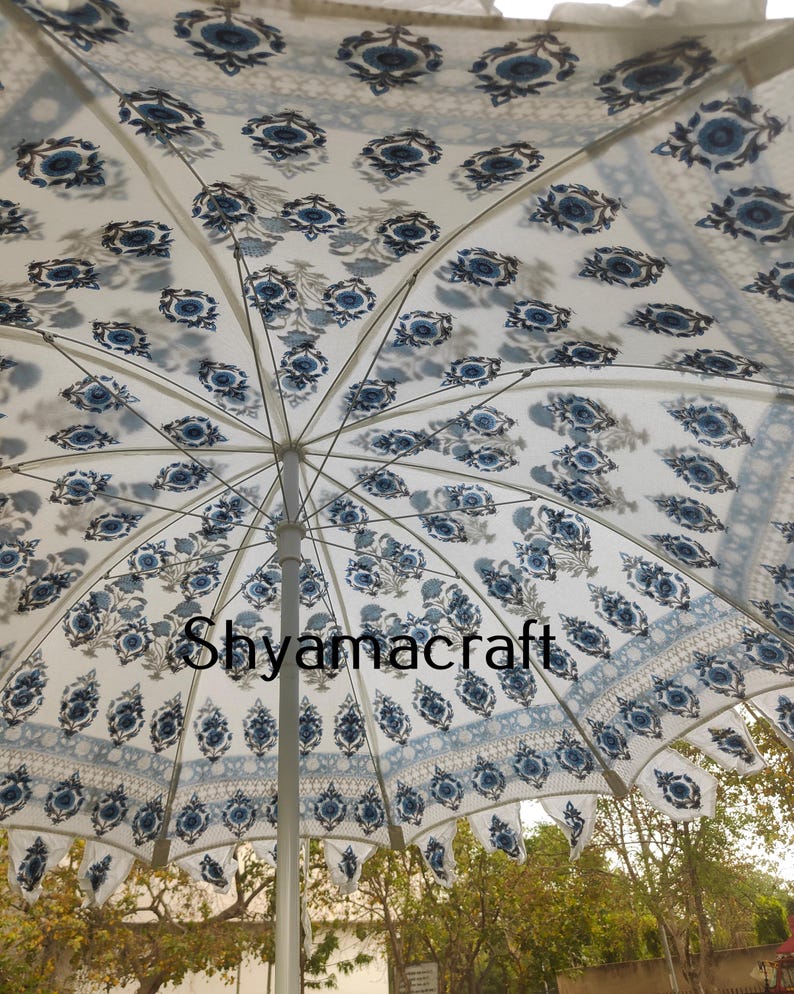 Puede incluir: Una sombrilla blanca y azul con un poste plateado. La sombrilla tiene un dise&ntilde;o floral con flores y hojas azules sobre un fondo blanco. La sombrilla est&aacute; abierta y la tela est&aacute; estirada.