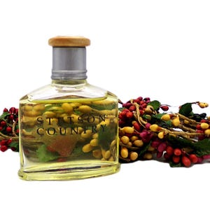Puede incluir: Una botella de vidrio transparente de colonia Stetson Country con tapa plateada y parte superior de madera. La botella est&aacute; llena de un l&iacute;quido amarillo. Una guirnalda decorativa de bayas rojas, amarillas y verdes est&aacute; al lado.