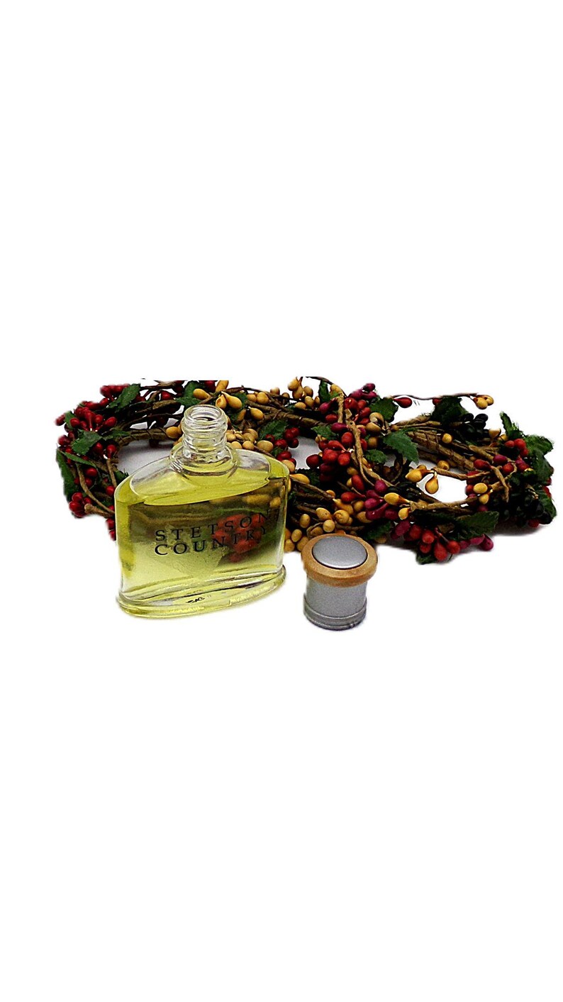 Puede incluir: Una botella de vidrio transparente de colonia Stetson Country con una tapa plateada y una corona de bayas decorativa. La botella est&aacute; llena de un l&iacute;quido amarillo. La corona presenta bayas rojas y amarillas con hojas verdes.