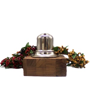 May include: A silver metal bottle stopper with a domed top sits on a wooden base. The base is made of small squares of wood and has a dark brown color. The bottle stopper is surrounded by a garland of red and yellow berries.