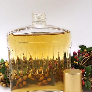 May include: A clear glass bottle with a gold cap, filled with a light amber liquid. The bottle has a textured surface and the word "Stetson" is etched on the front. The bottle is sitting on a white surface next to a gold cap.