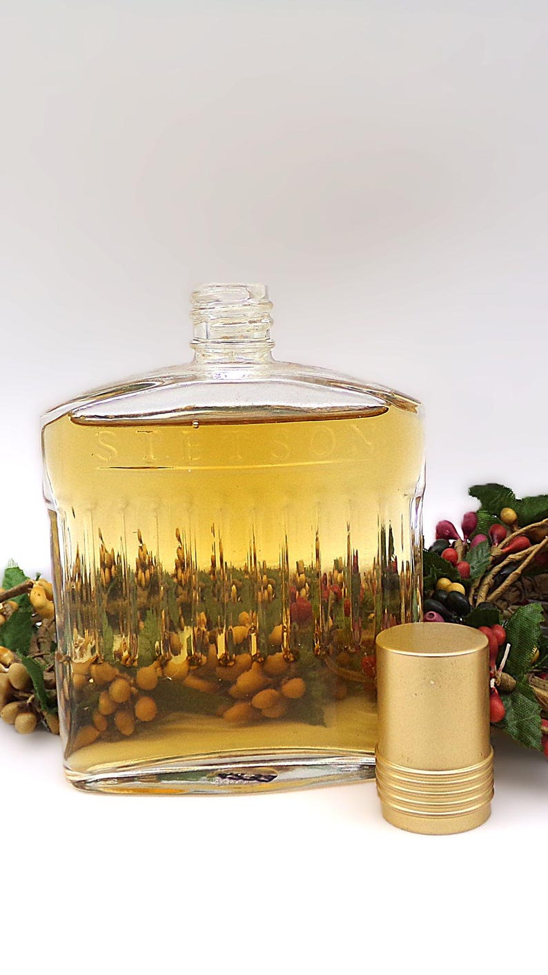 May include: A clear glass bottle with a gold cap, filled with a light amber liquid. The bottle has a textured surface and the word "Stetson" is etched on the front. The bottle is sitting on a white surface next to a gold cap.