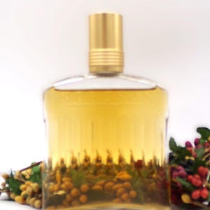 May include: A clear glass bottle with a gold cap filled with a pale yellow liquid. The bottle is sitting on a white surface with a small bunch of dried flowers in the background.