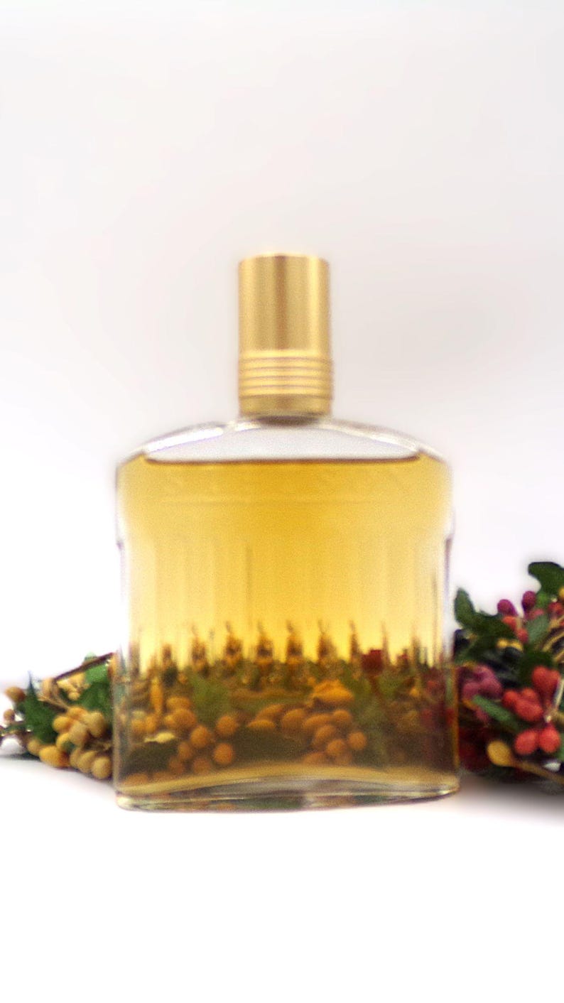 May include: A clear glass bottle with a gold cap filled with a pale yellow liquid. The bottle is sitting on a white surface with a small bunch of dried flowers in the background.