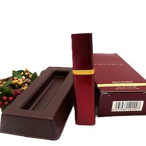 May include: A red perfume bottle with a gold band and a matching box. The box is dark red with the word "BOUCHERON" printed on it. A dark brown tray and a small floral arrangement are also in the image.