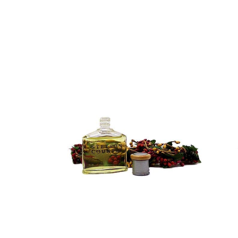 Puede incluir: Una botella de vidrio transparente con el texto "Stetson Country" est&aacute; llena de un l&iacute;quido amarillo. Una tapa plateada y marr&oacute;n se encuentra junto a la botella. Bayas rojas y amarillas est&aacute;n dispuestas alrededor de la botella.
