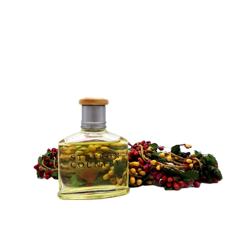 Puede incluir: Una botella de vidrio transparente de colonia Stetson Country con tapa plateada y parte superior de madera. La botella est&aacute; llena de un l&iacute;quido amarillo. Una guirnalda decorativa de bayas rojas, amarillas y verdes est&aacute; al lado.