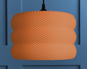 Puede incluir: Una lámpara colgante naranja con una superficie texturizada, similar a un patrón de panal. La lámpara tiene un diseño redondeado y en capas, y cuelga de un cable negro. El fondo es una pared azul.