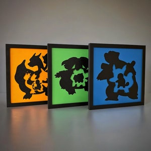 May include: Three framed wall art prints featuring black silhouettes of Pokemon characters on a colored background. The prints are in orange, green, and blue frames.