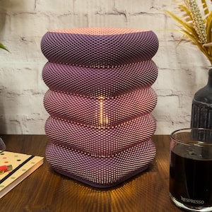 May include: A contemporary lamp with a textured design, featuring a colour gradient from peach to purple and pink. The lamp is stacked in a unique shape. It sits on a wooden surface, with a coffee glass and a notebook visible.