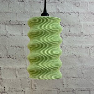 May include: A green and white patterned pendant light with a wavy, cylindrical design. The light hangs from a black cord and fixture. The textured surface of the light contrasts with the white brick wall in the background.