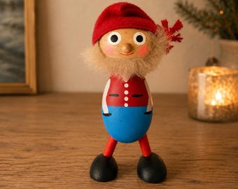 May include: A wooden figurine of a gnome with a red felt hat, a beige fuzzy beard, and a painted face. The body features a red vest with white buttons over a blue lower section. It has red legs and black rounded feet, standing upright.