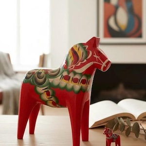 May include: A large, red wooden Dala horse with colourful floral patterns on its head and body. A smaller, matching horse is next to it. The traditional Swedish folk art piece is displayed on a wooden surface.