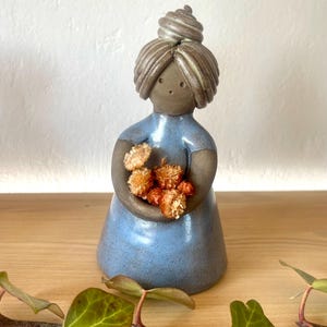 May include: A ceramic figurine of a woman in a blue dress, holding a small bouquet of dried orange flowers. The figurine has a bun hairstyle and a neutral skin tone. The figure is on a wooden surface.