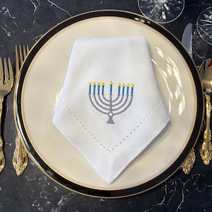 May include: White linen napkin with an embroidered menorah design, folded on a cream-colored plate with a black and gold rim. Gold silverware and a black marble surface complete the elegant table setting.