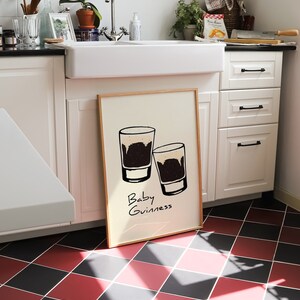 May include: A framed print with a white background and two black and white illustrations of glasses with the text "Baby Guinness" written below.