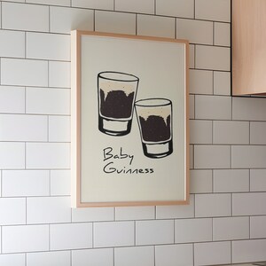 May include: A framed print with a white background and a black and white illustration of two shot glasses filled with a dark liquid. The text "Baby Guinness" is written below the glasses.
