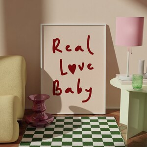 May include: A framed print with the text "Real Love Baby" in red lettering with a red heart replacing the "o" in "Love". The print is on a light pink background and is sitting on a green and white checkered rug.