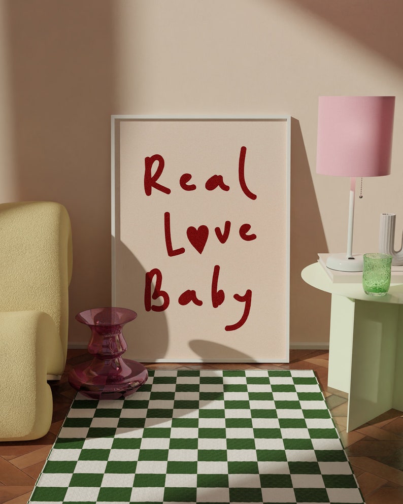 May include: A framed print with the text "Real Love Baby" in red lettering with a red heart replacing the "o" in "Love". The print is on a light pink background and is sitting on a green and white checkered rug.